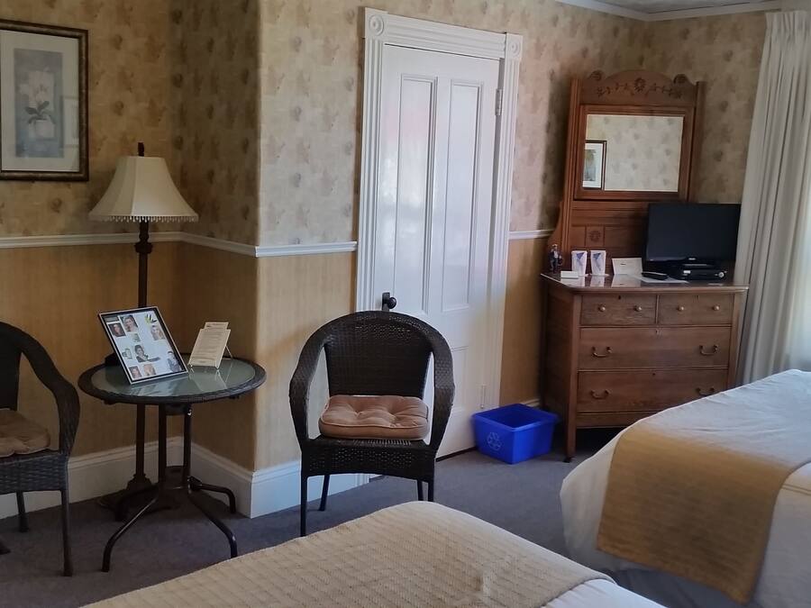 Habitación de hotel con dos camas individuales, una mesita con lámpara, una silla, un aparador con televisor y una ventana con vista a zonas verdes.