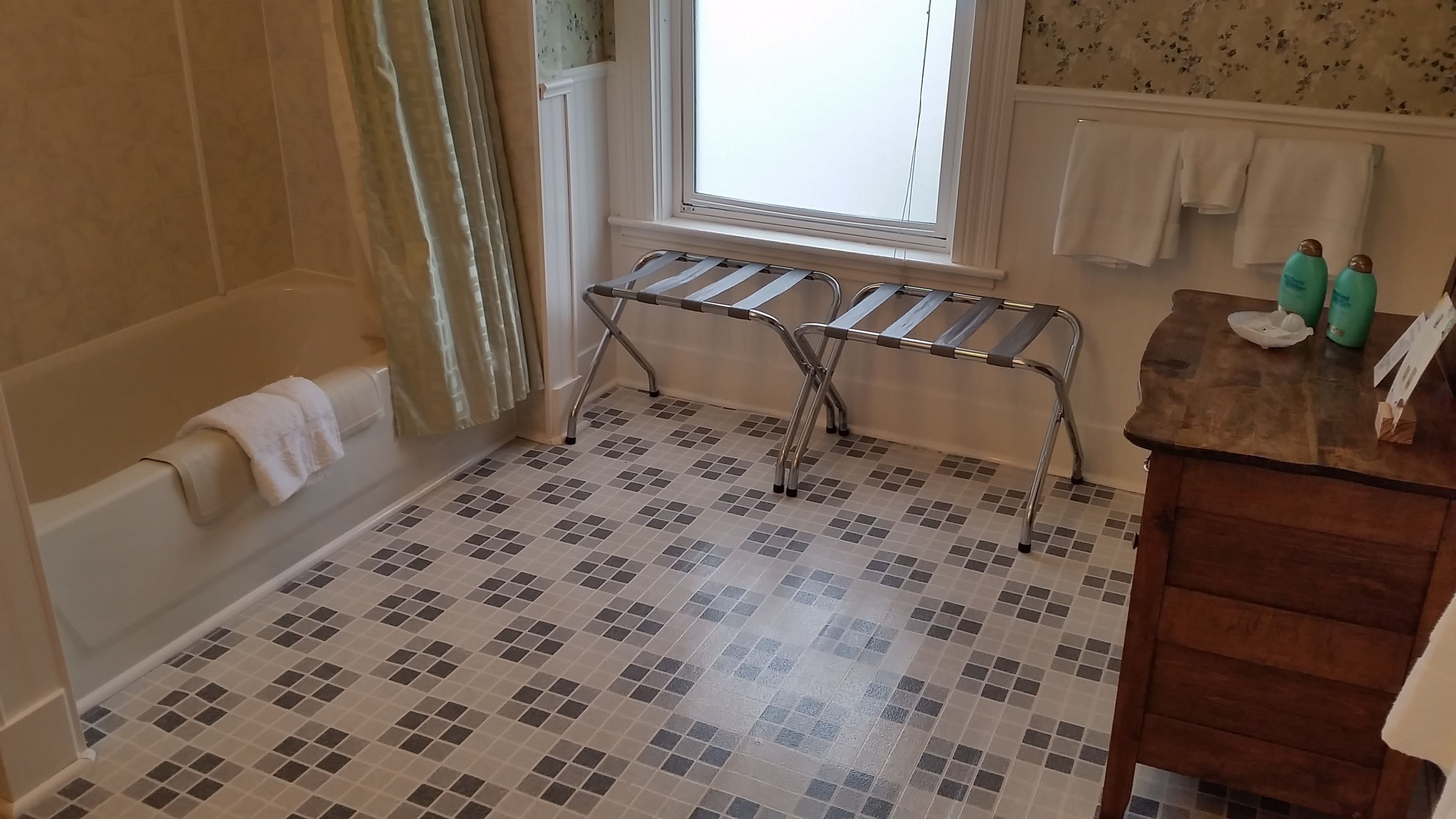 Un baño con suelo de baldosas, una bañera, una ventana con cortinas, un perchero para toallas y un tocador de madera con artículos de aseo.