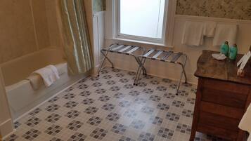 Un baño con suelo de baldosas, una bañera, una ventana con cortinas, un perchero para toallas y un tocador de madera con artículos de aseo.