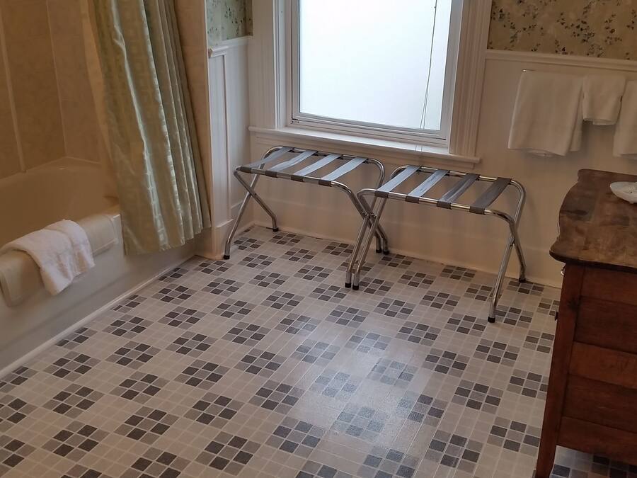 Un baño con suelo de baldosas, una bañera, una ventana con cortinas, un perchero para toallas y un tocador de madera con artículos de aseo.