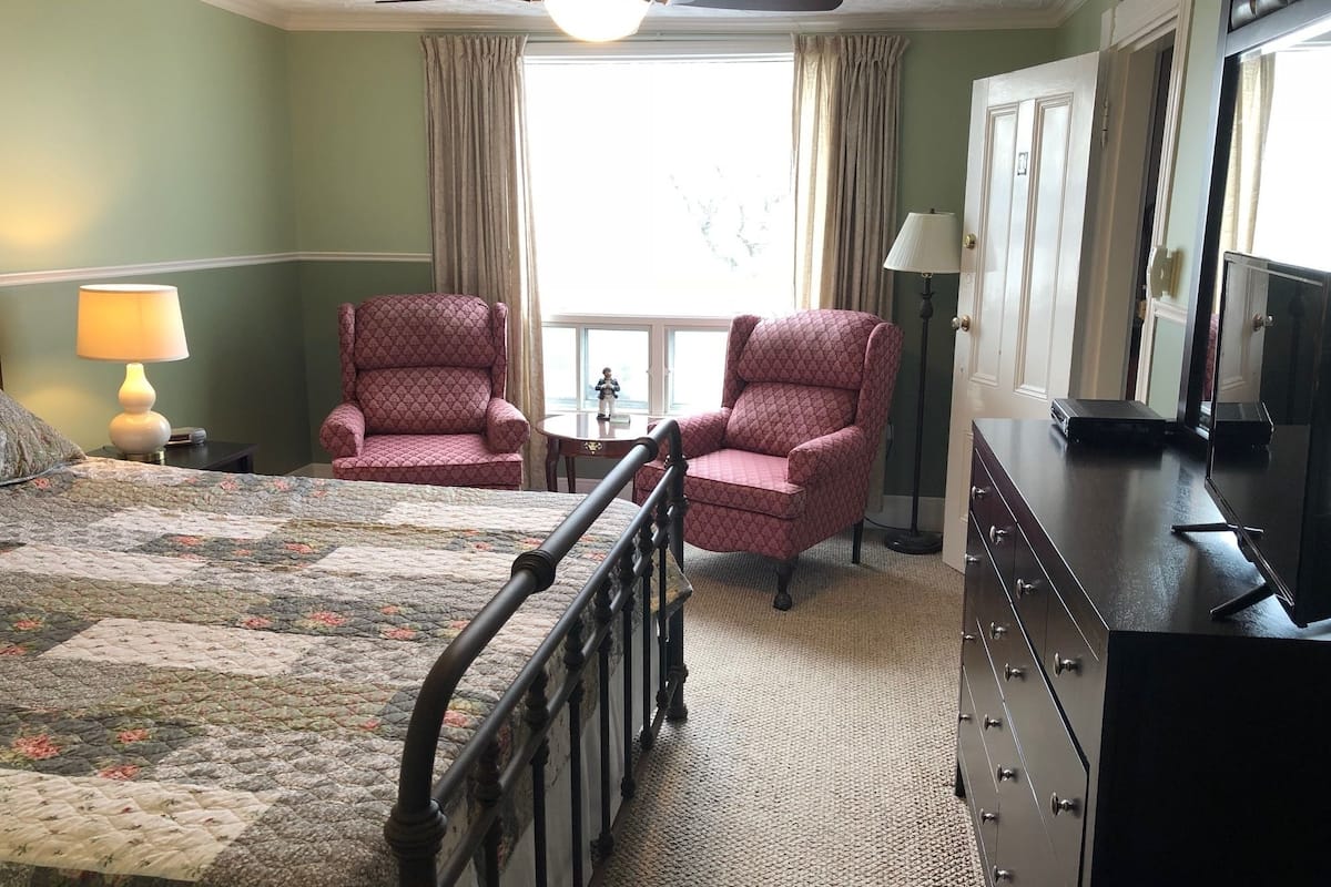 A hotel room with a bed, two armchairs, a dresser, a television, and a window with curtains.