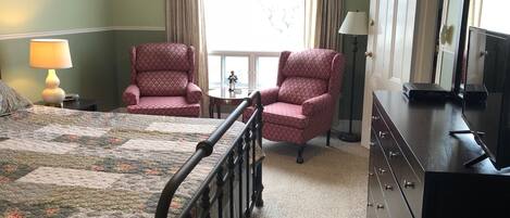 Une chambre d’hôtel comprenant un lit, deux fauteuils, une commode, un téléviseur et une fenêtre avec des rideaux.