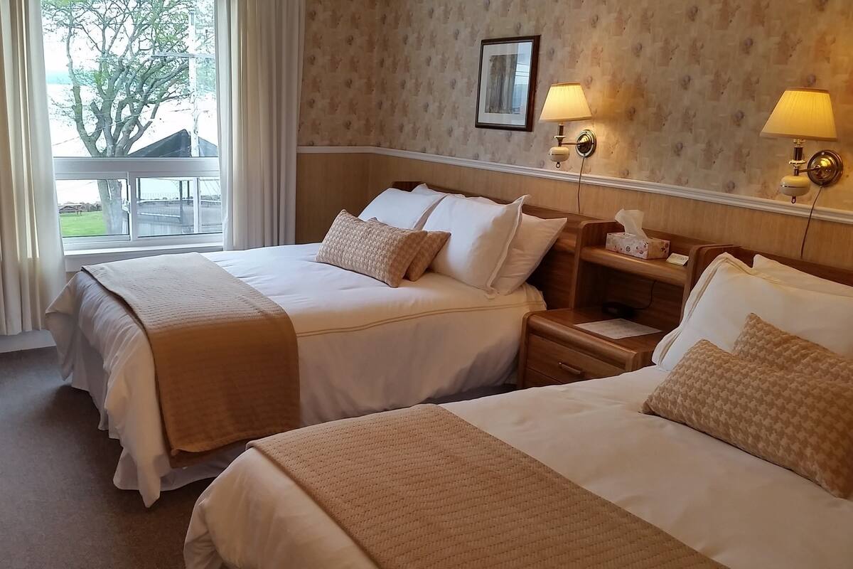 A hotel room with two beds, a TV, a bedside table with a lamp, and a view of the outdoors.