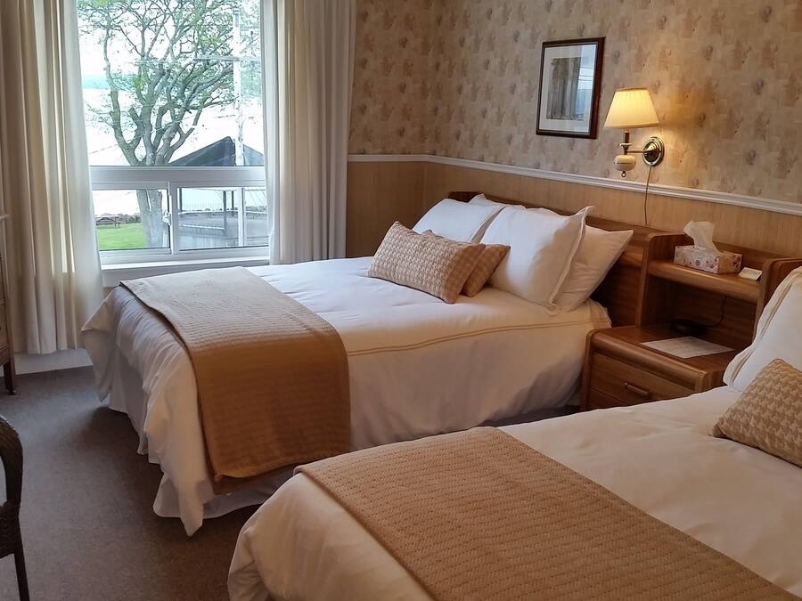 Quarto de hotel com duas camas, televisão, mesa de cabeceira com luminária e vista para o exterior.