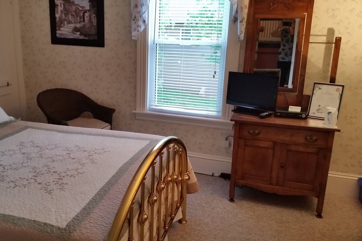 A bedroom with a bed, a chair, a dresser, a television, and a window with blinds.