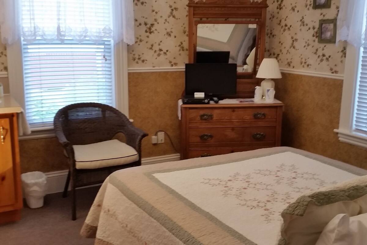 A bedroom with a bed, a dresser, a chair, a mirror, and a window with blinds.