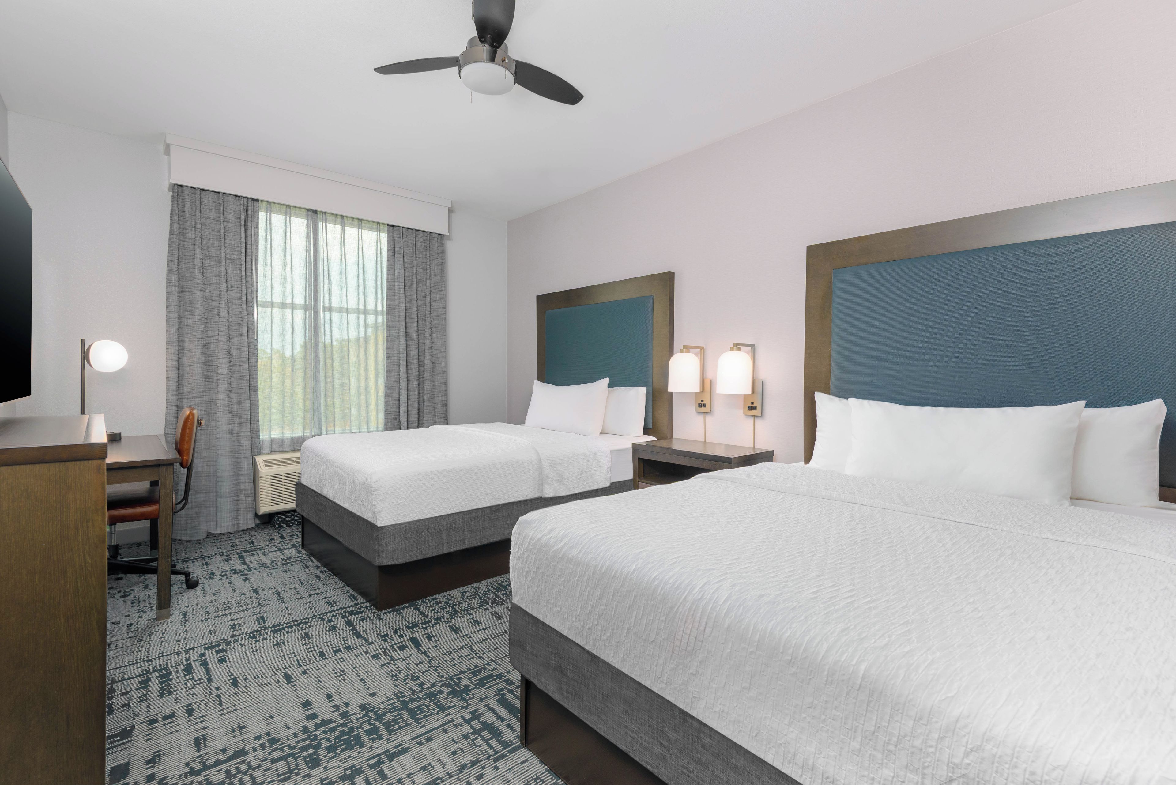 A hotel room with two beds, a desk, a chair, a ceiling fan, and a large window with curtains.