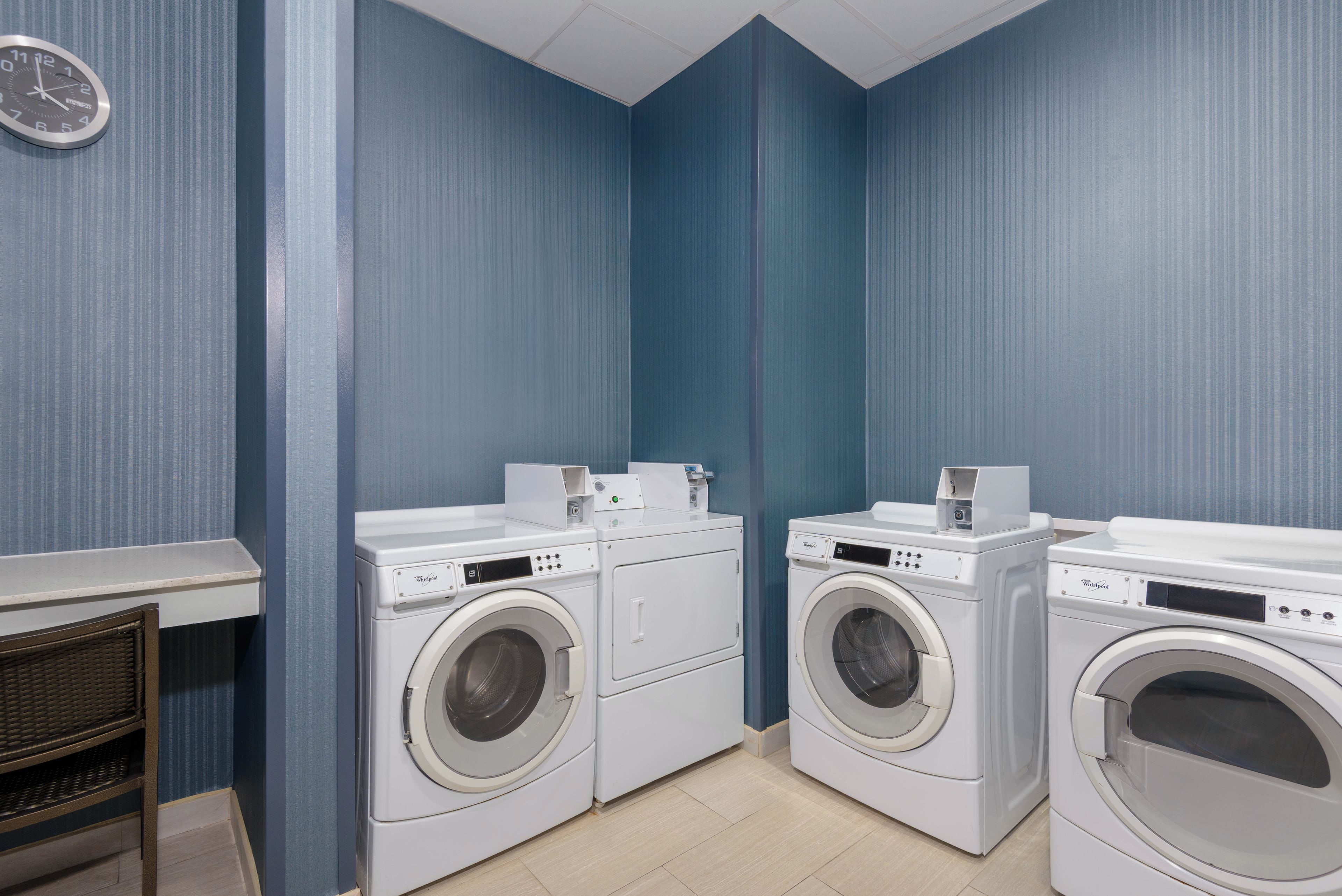 A laundry room with two front-loading washing machines, a small table, and a wicker chair.