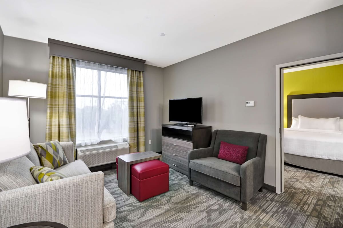 A hotel room with a grey sofa, a red ottoman, a television, a wardrobe, and a bed with a yellow accent wall.