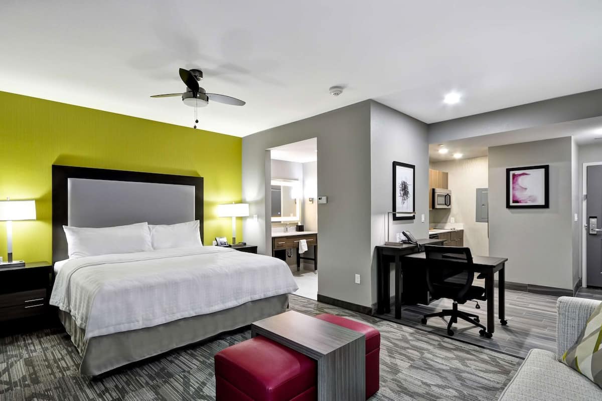 A hotel room with a bed, a desk, a chair, a red ottoman, a ceiling fan, and a bathroom visible through an open door.