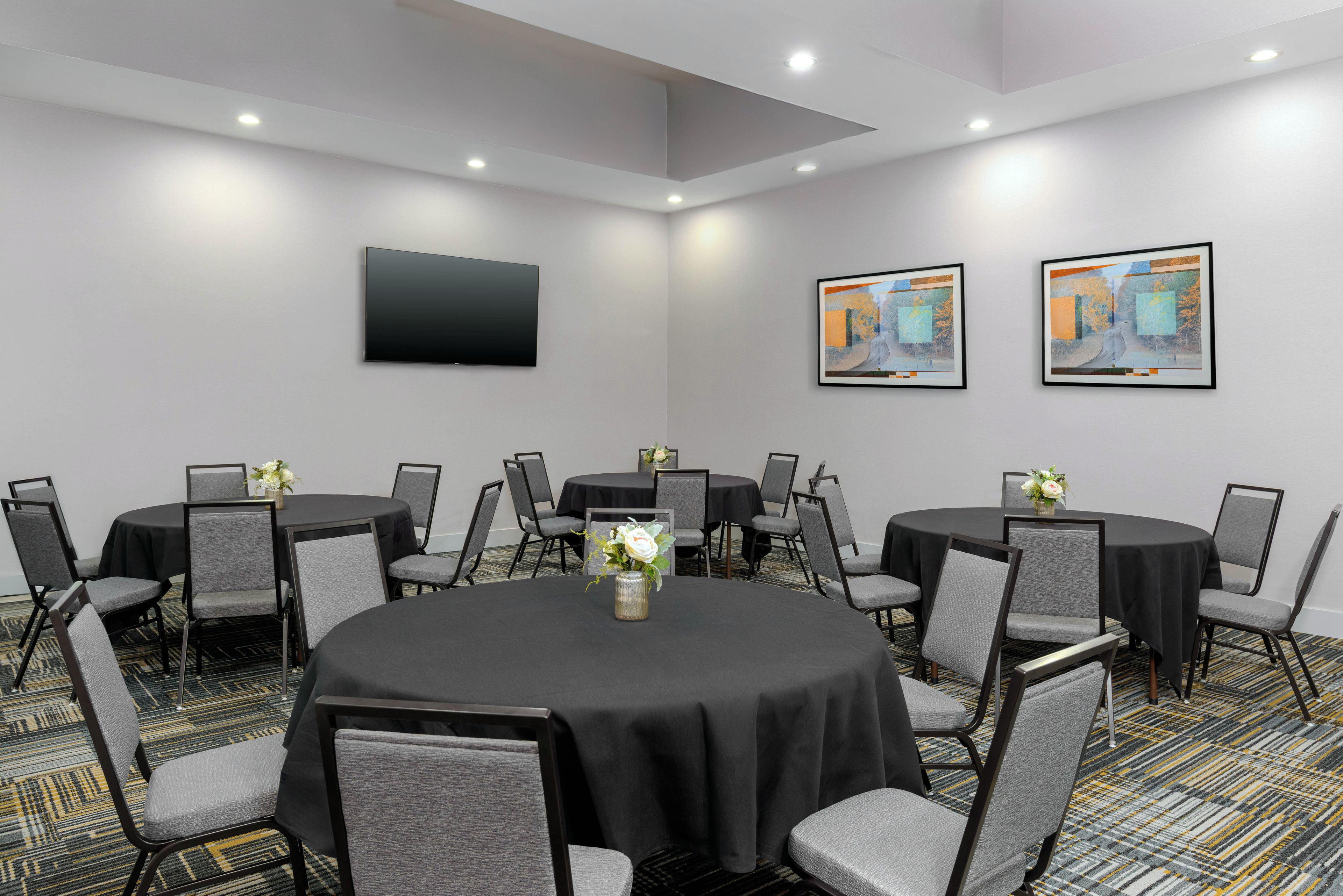 A conference room with round tables, grey chairs, a flat-screen TV, and two framed artworks on the wall.