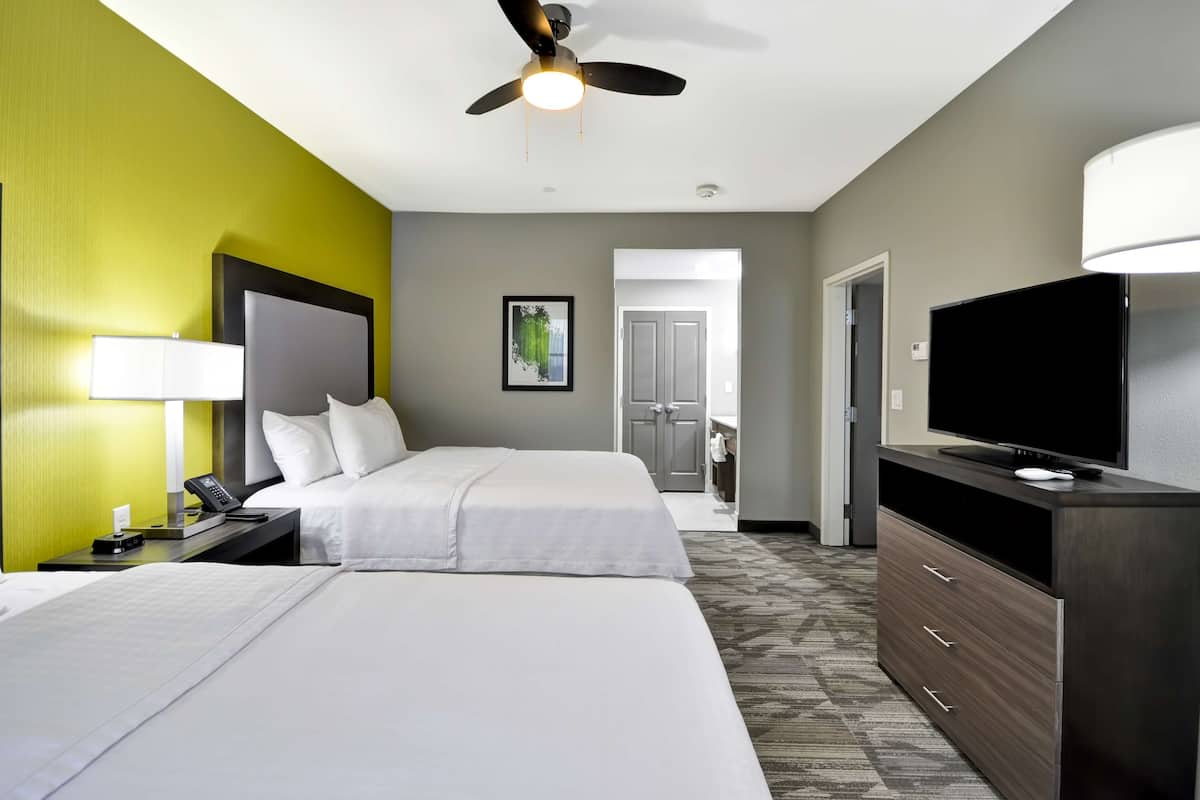 A hotel room with two beds, a desk with a lamp, a television, and a ceiling fan.
