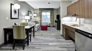 A modern hotel room with a dining area, a sofa, a red ottoman, a kitchen with a dishwasher, and a large window with curtains. - Homewood Suites by Hilton Tyler (Tyler)
