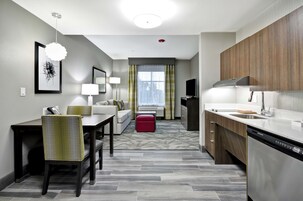 Una habitación de hotel moderna con comedor, un sofá, un reposapiés rojo, una cocina con lavavajillas y un gran ventanal con cortinas.