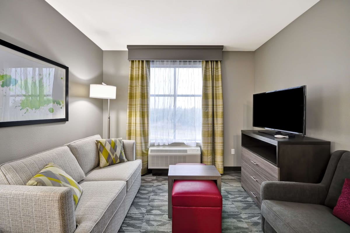 A hotel room with a sofa, a red ottoman, a TV on a wooden stand, and a window with curtains.