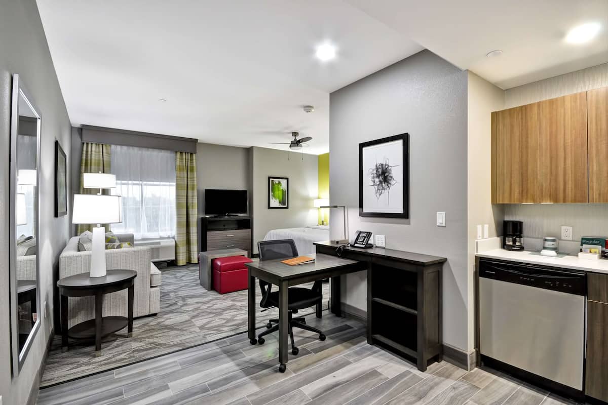 A compact hotel room with a kitchenette, a dining area, a sofa, a small table, and a red ottoman.