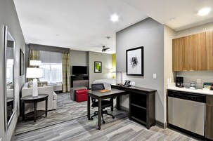 Habitación de hotel pequeña y funcional con kitchenette, zona de comedor, sofá, una mesita y un reposapiés rojo.