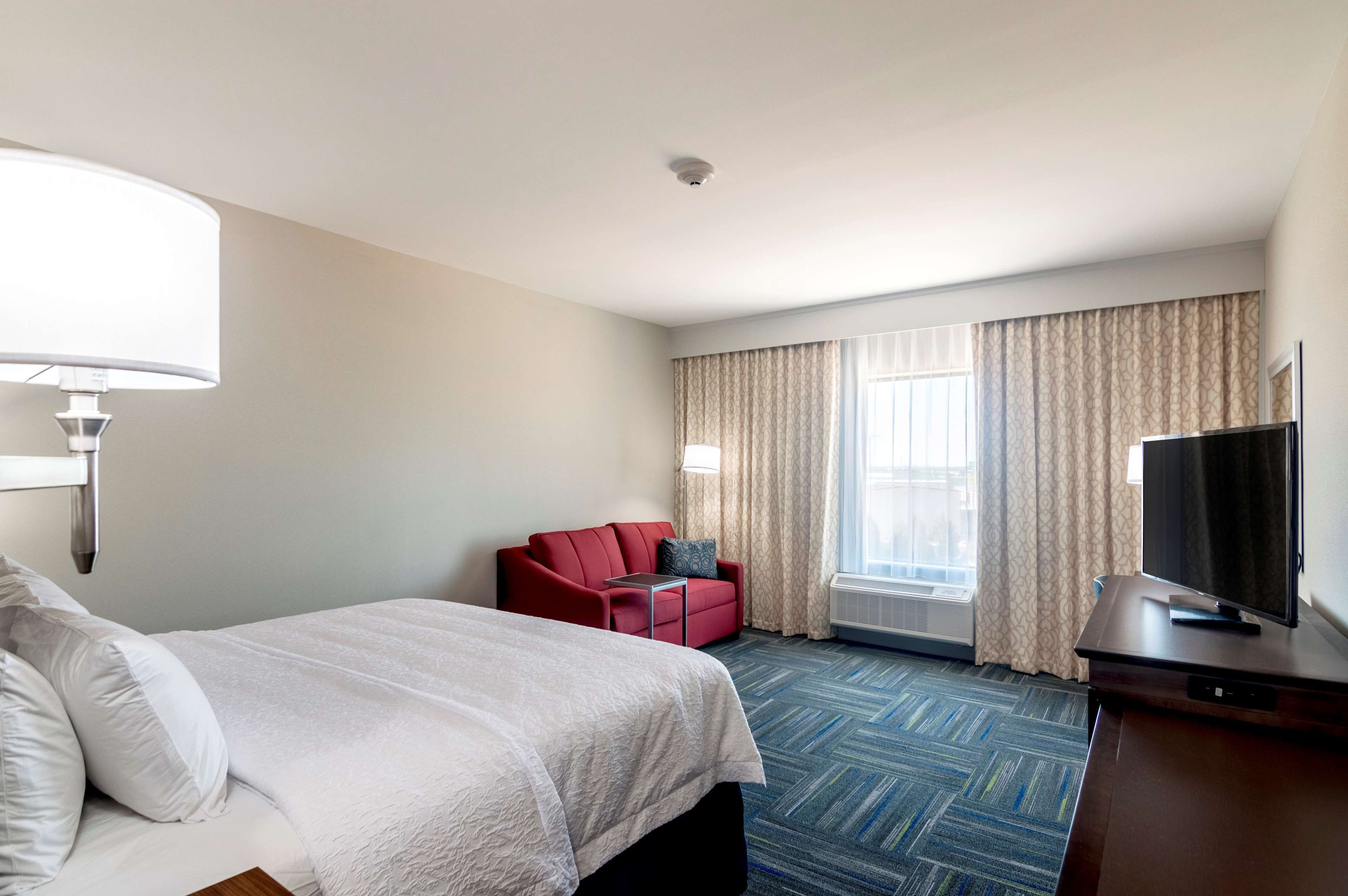 A hotel room with a bed, a red armchair, a television, and a large window with curtains.
