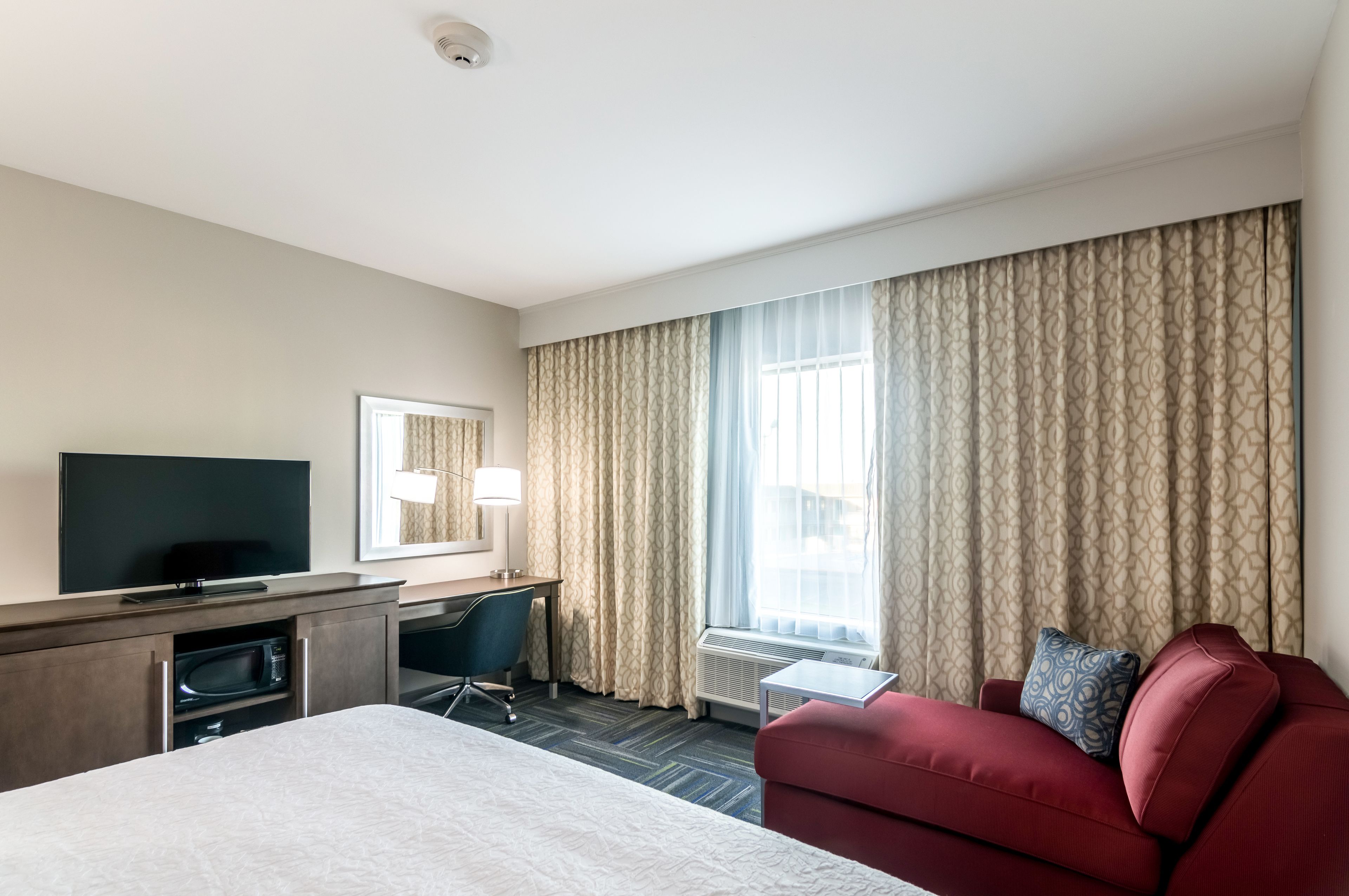 Une chambre d’hôtel avec un lit, un bureau avec un ordinateur, une télévision, un canapé rouge et une grande fenêtre avec des rideaux.