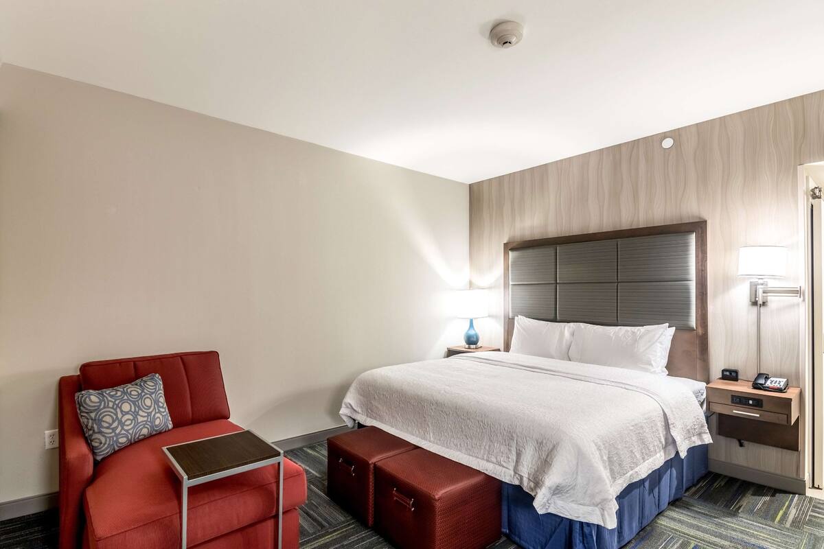 A hotel room with a bed, a red sofa, a small table, and a bedside table with a lamp and phone.