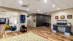 A modern hotel lobby with a wooden floor, a reception desk, a coffee station, and a seating area with a patterned rug.