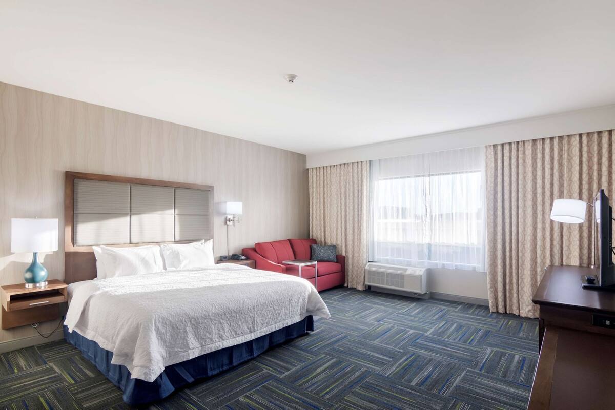 A hotel room with a bed, a red armchair, a desk with a lamp, and a large window with curtains.