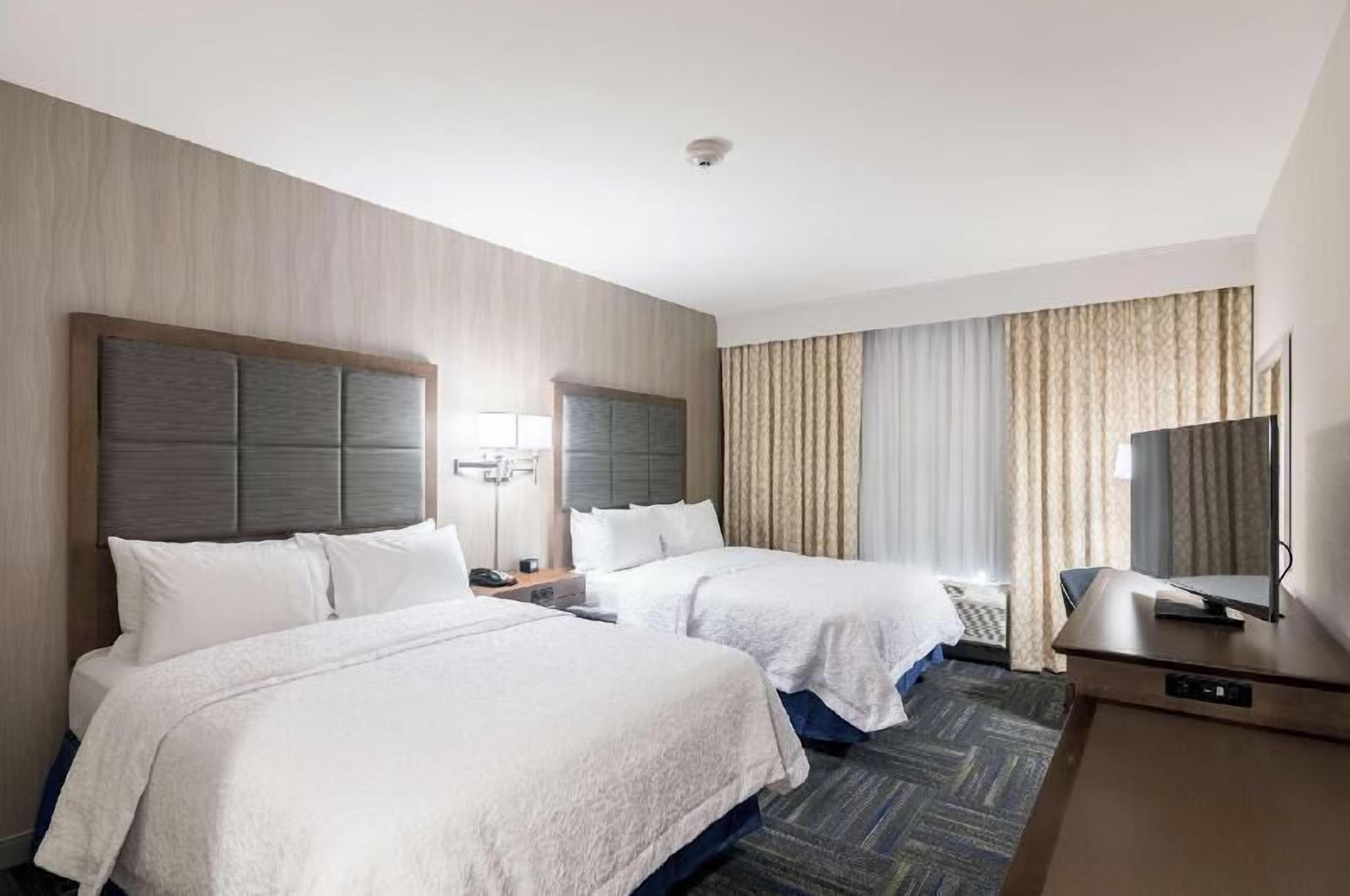 Habitación de hotel con dos camas, un escritorio, televisor y una ventana con cortinas.