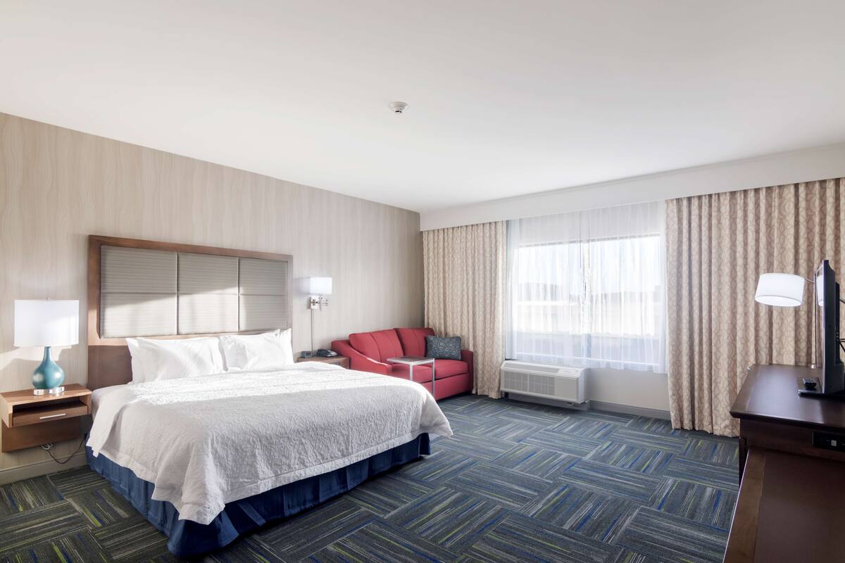 A hotel room with a large bed, a red armchair, a desk with a lamp, and a large window with curtains.