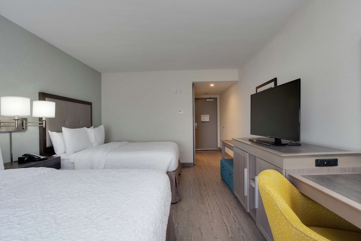 A hotel room with two beds, a desk with a television, a chair, and a nightstand with lamps.