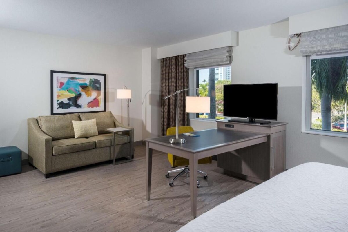 A hotel room with a bed, a desk, a television, a sofa, a lamp, and a window with a view.