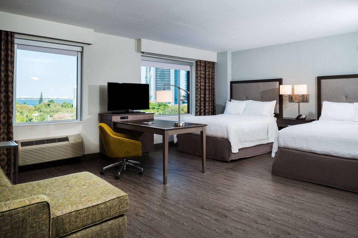 A hotel room with a desk, two beds, a television, and a view of the city.