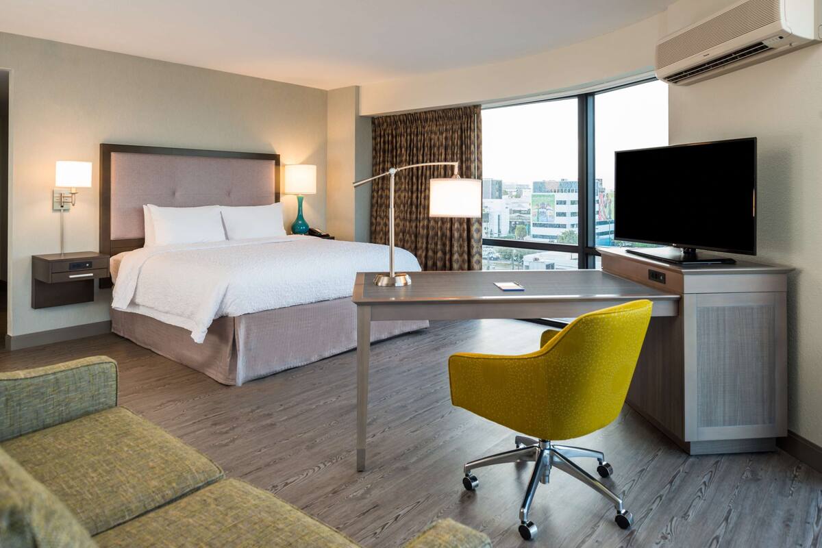 A hotel room with a bed, desk, chair, television, and a view of the city.