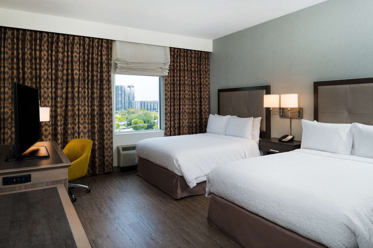 A hotel room with two beds, a desk with a TV, a chair, and a window with curtains.
