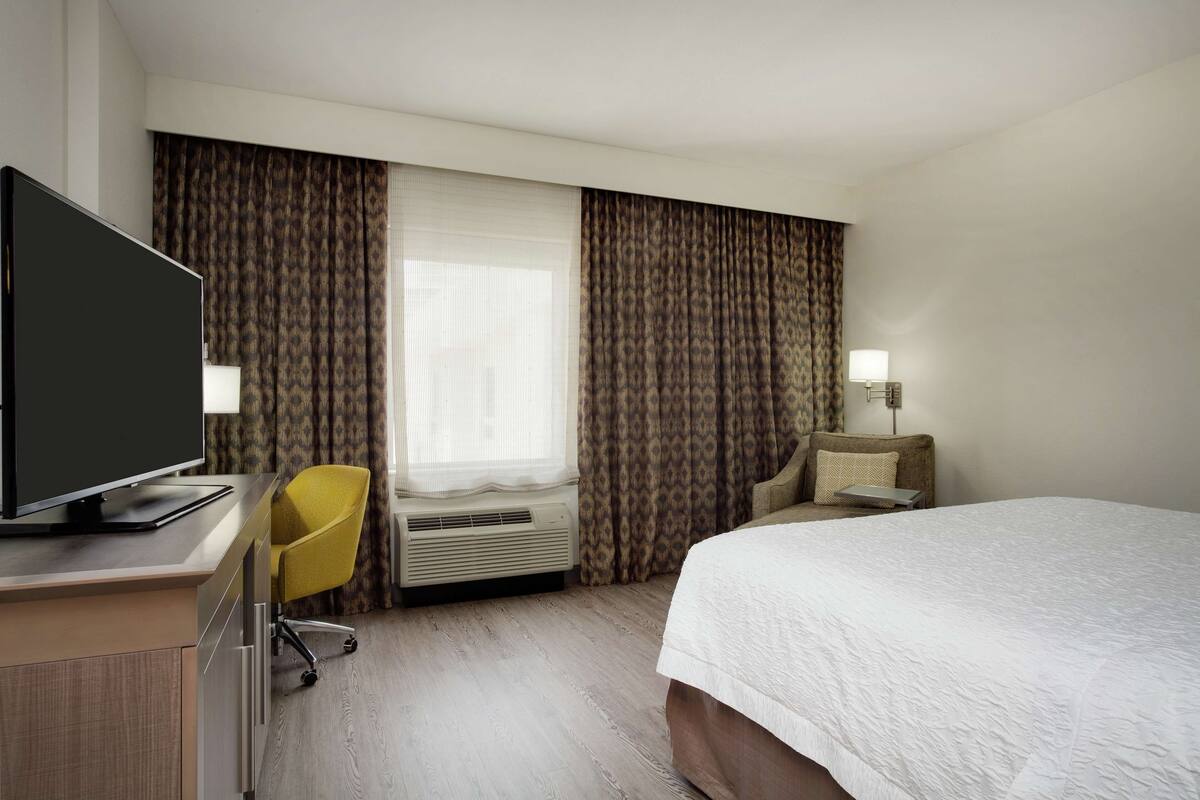 A hotel room with a bed, a desk with a television, a chair, a bedside table with a lamp, and a window with curtains.