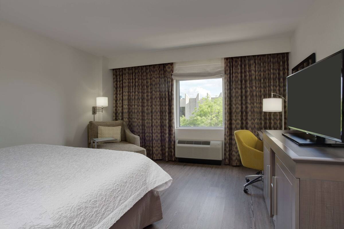 A hotel room with a large bed, a desk with a flat-screen TV, a chair, and a window with curtains.