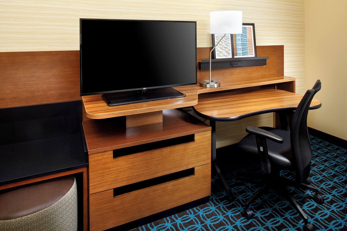 A hotel room with a wooden desk, a black chair, a flat-screen TV, a lamp, and a framed picture on the wall.
