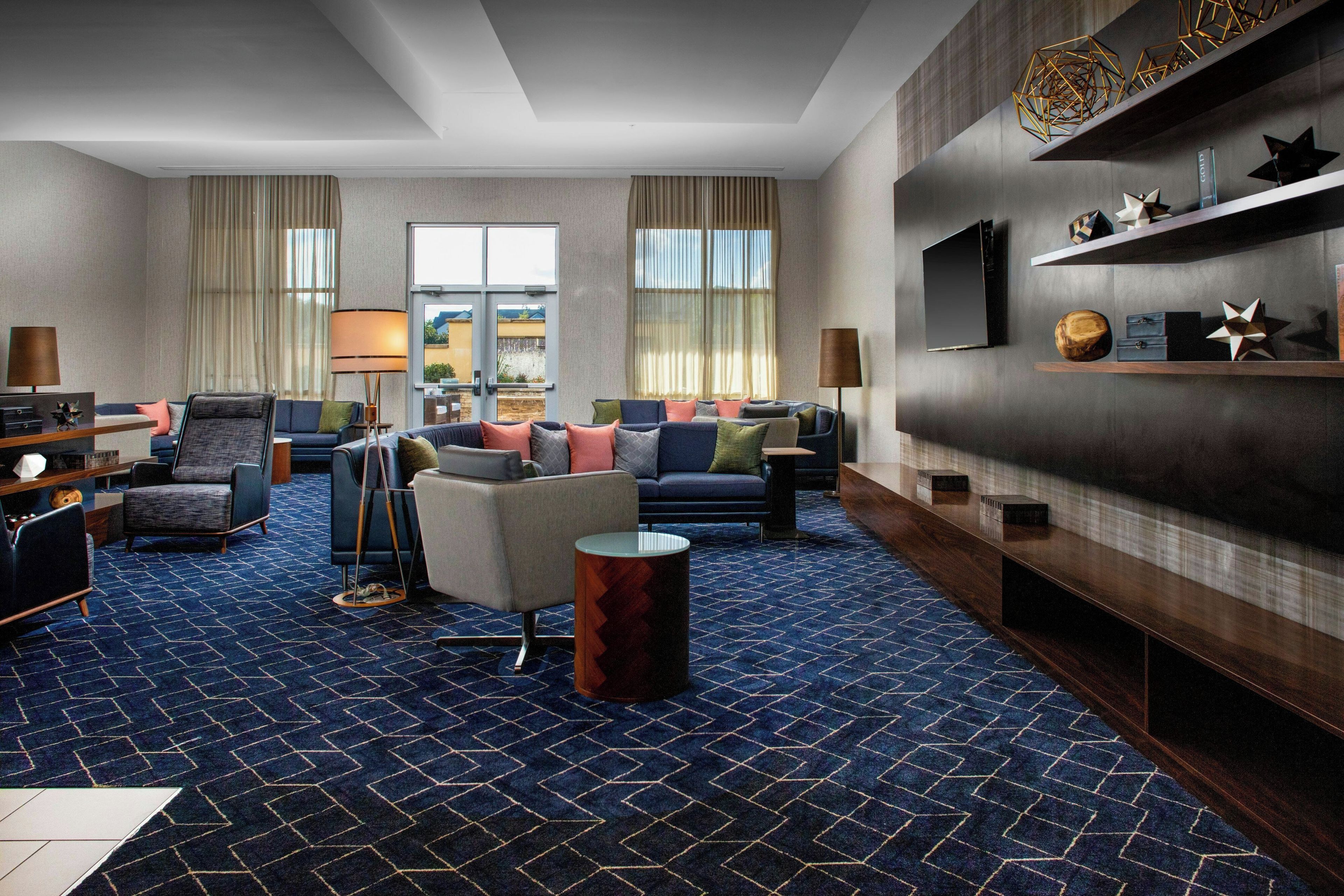A modern hotel room with a blue patterned carpet, a seating area with sofas and armchairs, a flat-screen TV mounted on the wall, and a wooden sideboard with decorative items.