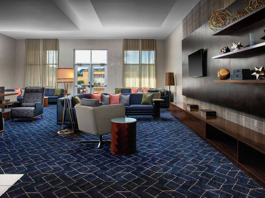 A modern hotel room with a blue patterned carpet, a seating area with sofas and armchairs, a flat-screen TV mounted on the wall, and a wooden sideboard with decorative items.