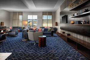 A modern hotel room with a blue patterned carpet, a seating area with sofas and armchairs, a flat-screen TV mounted on the wall, and a wooden sideboard with decorative items.