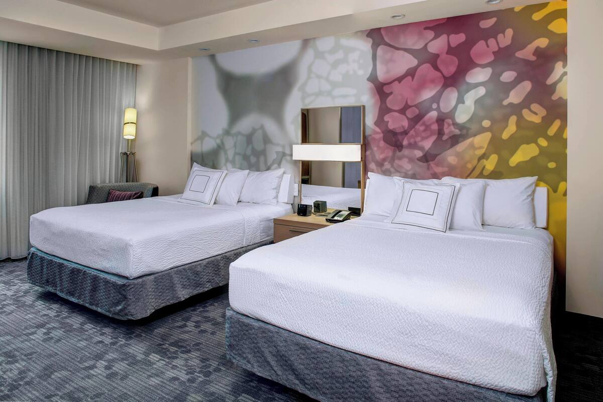 A hotel room with two beds, a desk, a lamp, and a large abstract wall mural.