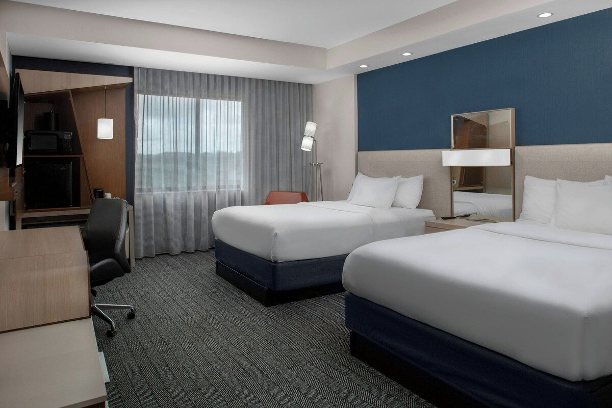 A hotel room with two beds, a desk, a chair, a wardrobe, and a large window with curtains.