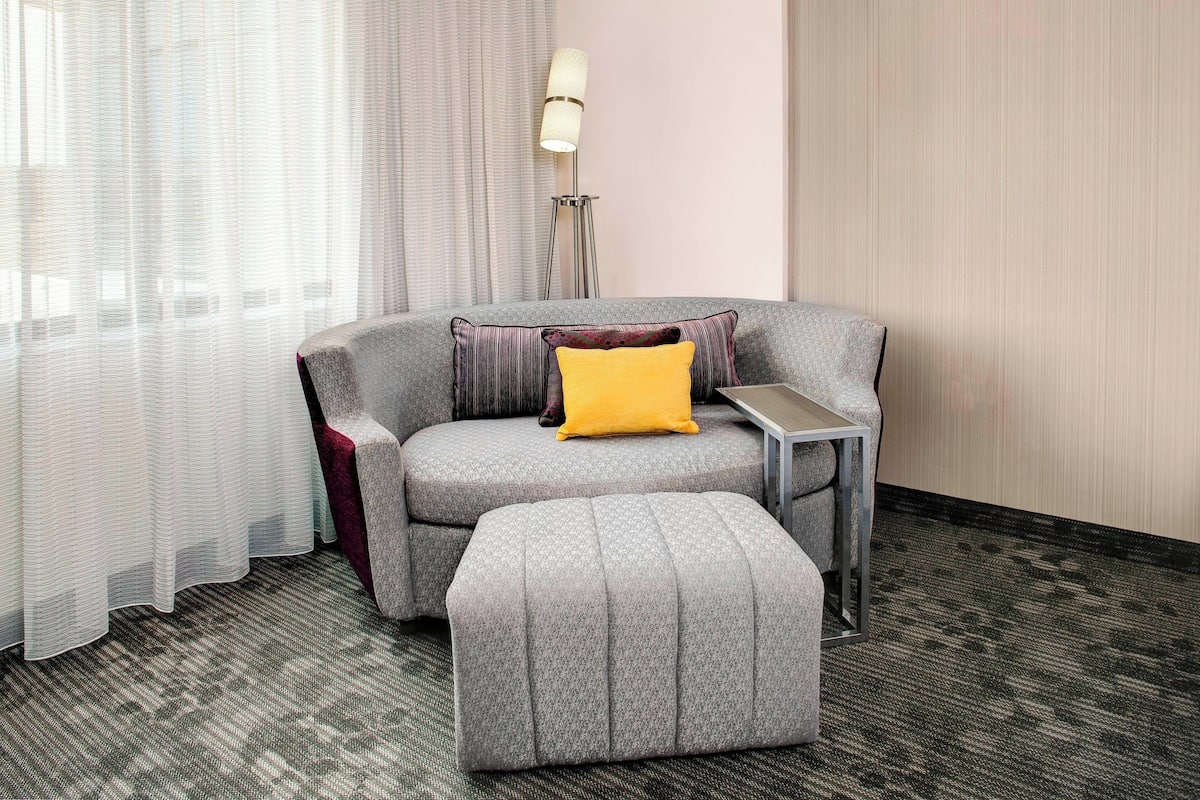 A modern hotel room with a grey sofa, a matching ottoman, a small side table, and a floor lamp.