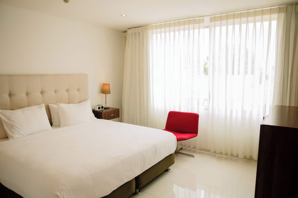 Habitación de hotel con una cama grande, una silla roja, una lámpara y vistas al exterior a través de cortinas translúcidas.