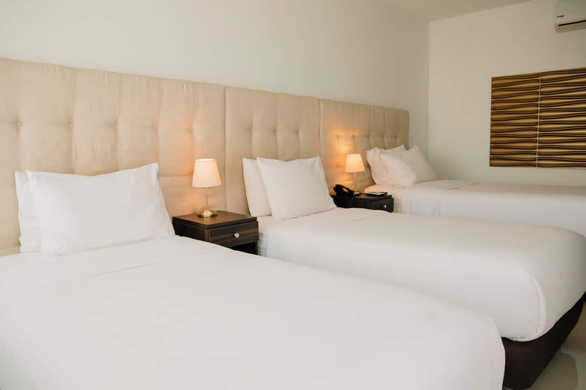 Habitación de hotel con tres camas, cada una con ropa de cama blanca y cabeceros de madera. Se observan mesitas de noche con lámparas y un aplique de pared.