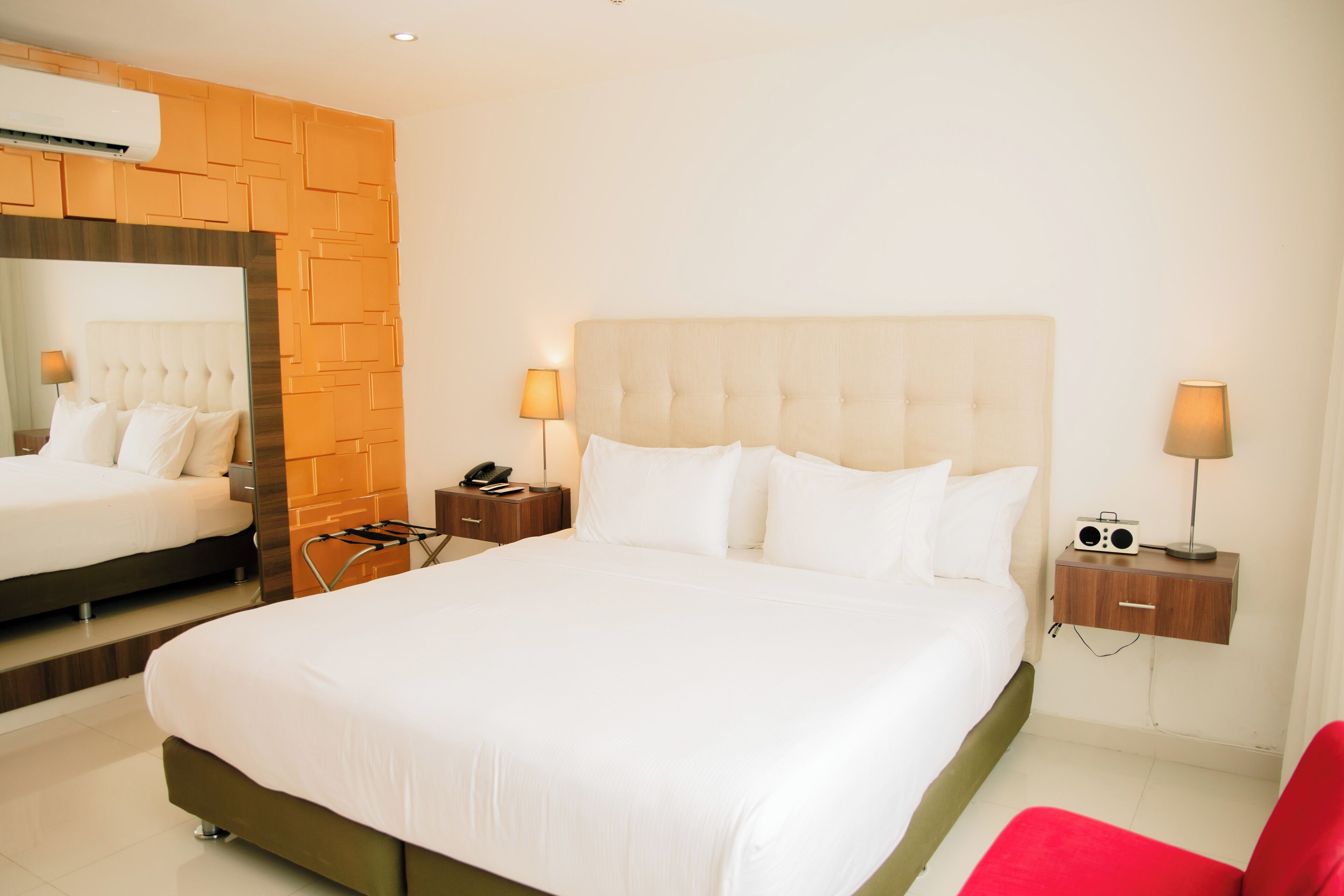 A hotel room with a large bed, a mirror, a nightstand with a lamp, and a red chair.