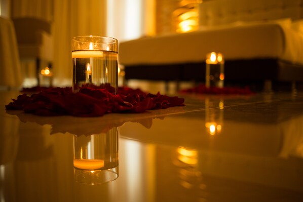 A candle in a glass holder on a reflective surface with rose petals, surrounded by candles and a softly lit background.