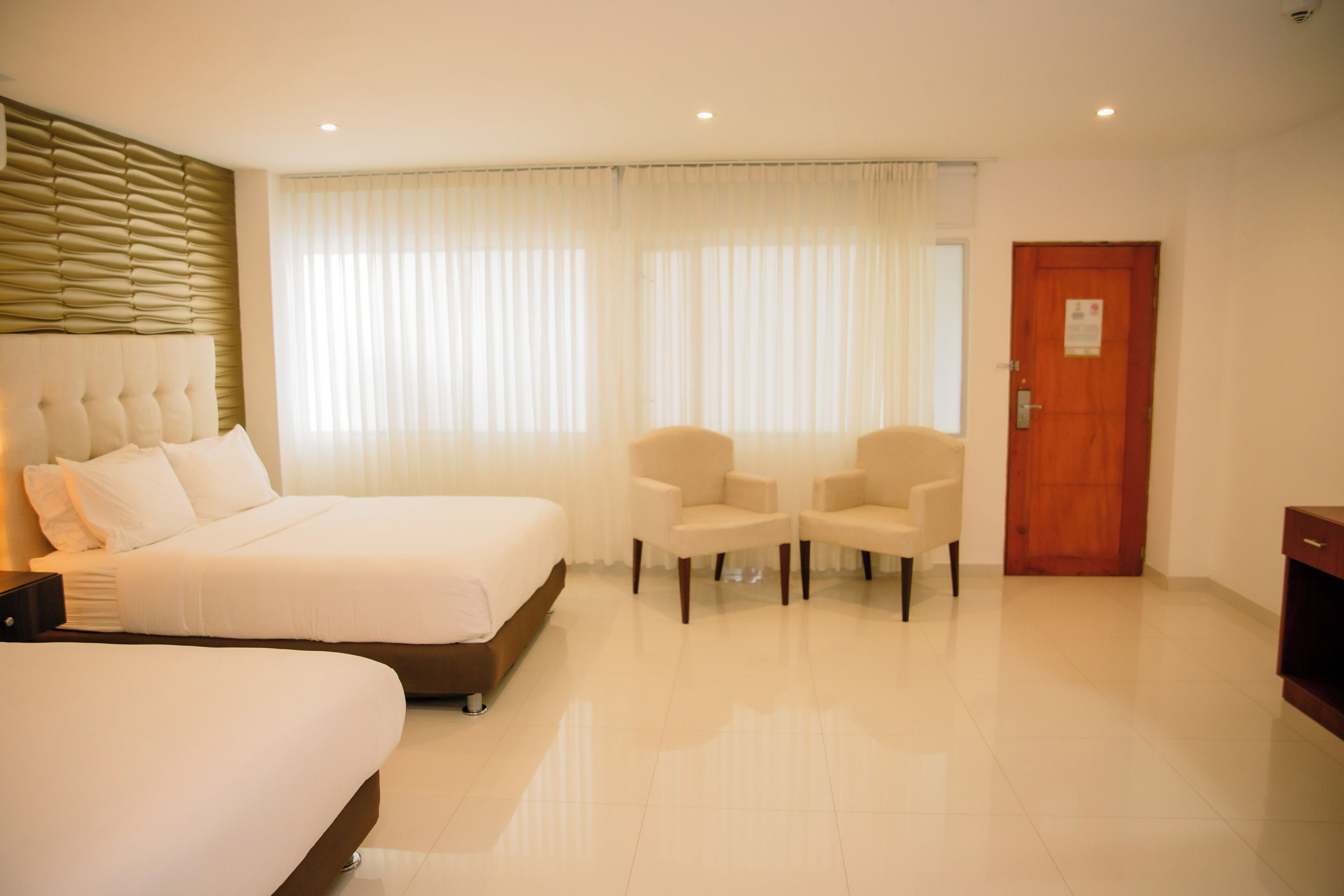 A hotel room with a large bed, two armchairs, a wooden door, and a window with sheer curtains.