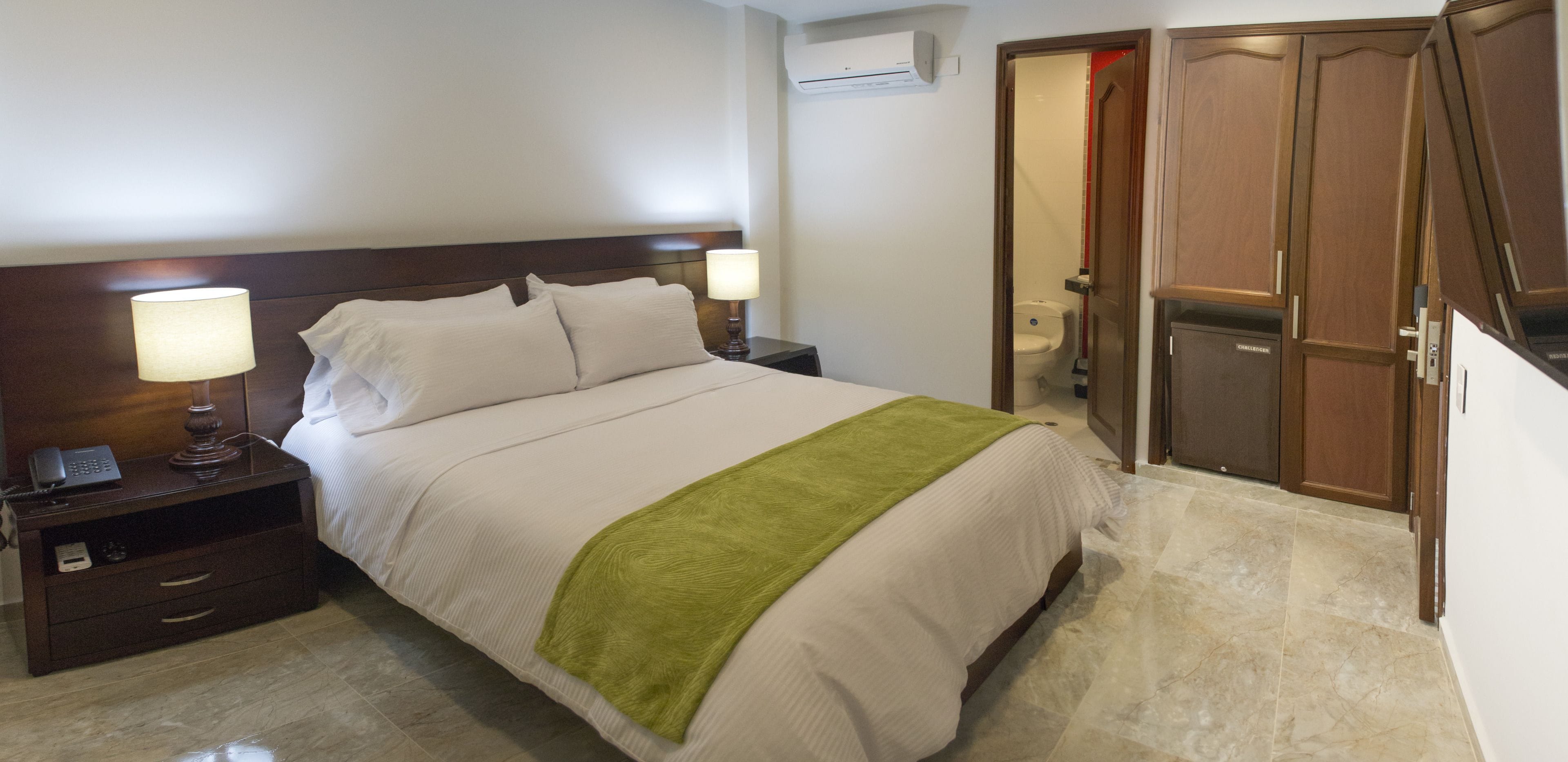 A hotel room with a bed, bedside lamps, a nightstand, a green throw blanket, a wooden wardrobe, and a toilet visible through an open door.