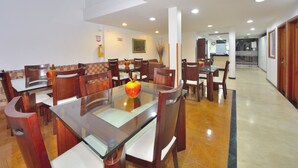 Un área de comedor con mesas de vidrio y sillas de madera, un florero con manzanas, y un cuadro en la pared.