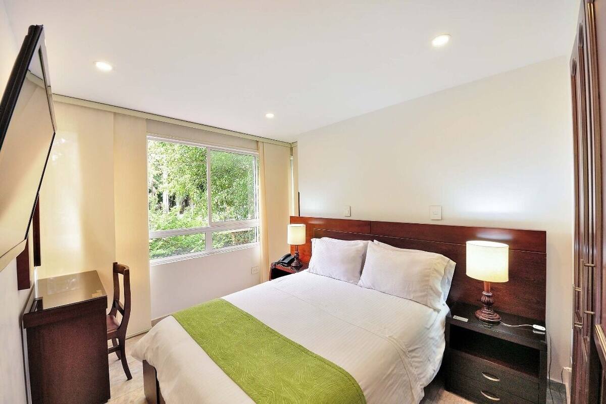A hotel room with a bed, bedside tables, a desk, a chair, a window with a view of greenery, and a TV mounted on the wall.